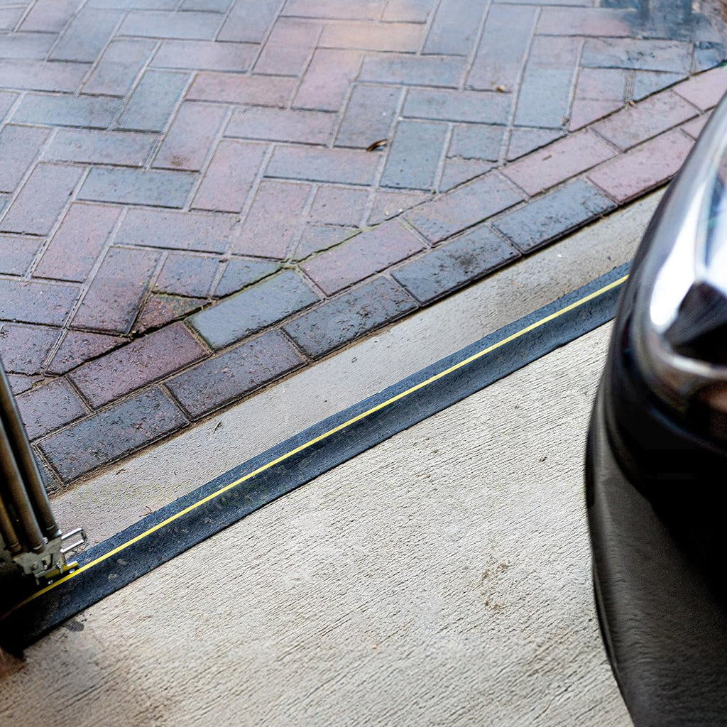 A close-up of a black rubber garage door seal strip with a yellow stripe, installed along the threshold next to a concrete driveway and brick pavers, with part of a black car visible on the right.